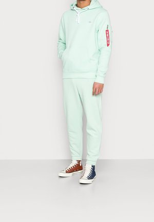 Felpa verde menta con lacci bianchi, caratterizzata da una tasca frontale e dettagli rossi sulle maniche. Abbinata a pantaloni coordinati e sneakers bicolore.