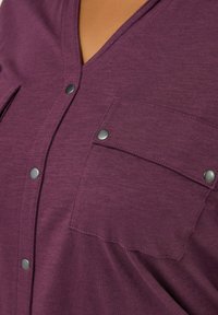 Top a V con scollo a V in morbido cotone misto di colore viola scuro, dotato di bottoni a pressione e una tasca sul petto con pattina curva. Texture liscia e design informale.