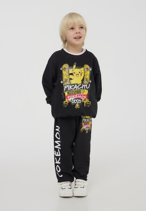Felpa nera di cotone con grafica di Pikachu e dettagli gialli, abbinata a pantaloni della tuta neri con testo "POKÉMON". Sneakers bianche.