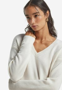 Weißer V-Ausschnitt-Pullover mit gerippten Bündchen und subtiler Struktur, lässig getragen. Das weiche Strickmaterial verleiht einen entspannten Look.