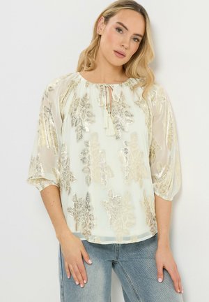 Blondynka w kremowej bluzce z metalicznymi złotymi wzorami kwiatowymi i troczkami, w zestawieniu z jasnoniebieskimi dżinsami, stojąca na tle białej ściany.