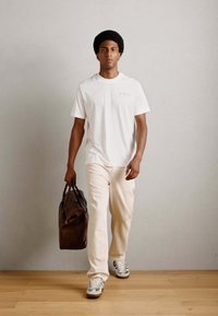 White crewneck t-shirt, beige pants, and gray sneakers. Brown leather duffle bag in hand. Simple, functional design. Indoor setting.