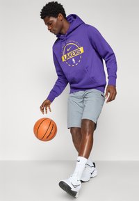 Purple hoodie with yellow "Lakers" logo, gray shorts, basketball, and white sneakers. Features a front pocket and drawstring hood.