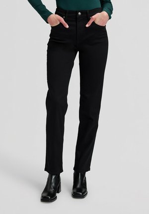 Person trägt schwarze Jeans mit geradem Bein, schwarze Knöchelstiefel und ein dunkelgrünes langärmeliges Oberteil mit Händen in den Taschen.