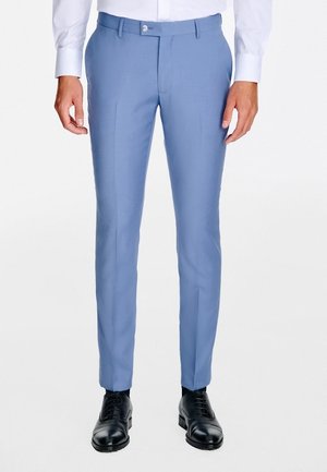 Mężczyzna noszący dopasowane jasnoniebieskie spodnie garniturowe, czarne eleganckie buty i białą koszulę z długim rękawem, stojący na białym tle.