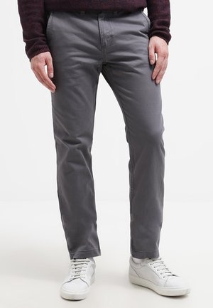 Mand iført grå bukser og hvide sneakers, står med hænderne afslappet ned langs siden på hvid baggrund.