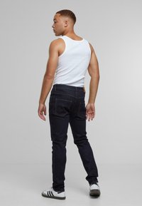 Sötétkék, slim fit farmernadrág bőr címkével a derékpánton, fehér trikóval és fehér sportcipővel viselve.