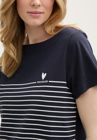 Camiseta de algodón de color azul marino con mangas cortas, que presenta franjas horizontales blancas y un logotipo de corazón encima del nombre de la marca "TOM TAILOR."