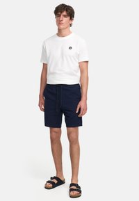 Biała bawełniana koszulka z czarnym logo na piersi, zestawiona z fakturowanymi granatowymi szortami i czarnymi sandałami z regulowanymi paskami.