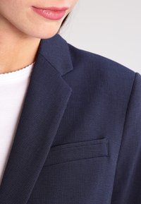 Marineblauer Blazer aus strukturiertem Stoff, mit Reverskragen und einer einzelnen Brusttasche, getragen über einem weißen Hemd.