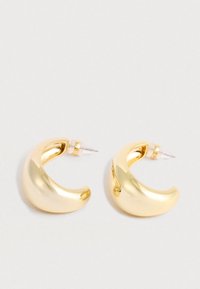 Une paire de créoles en or brillant et épais en forme de croissant, avec des fermoirs transparents à pression sur un fond blanc.