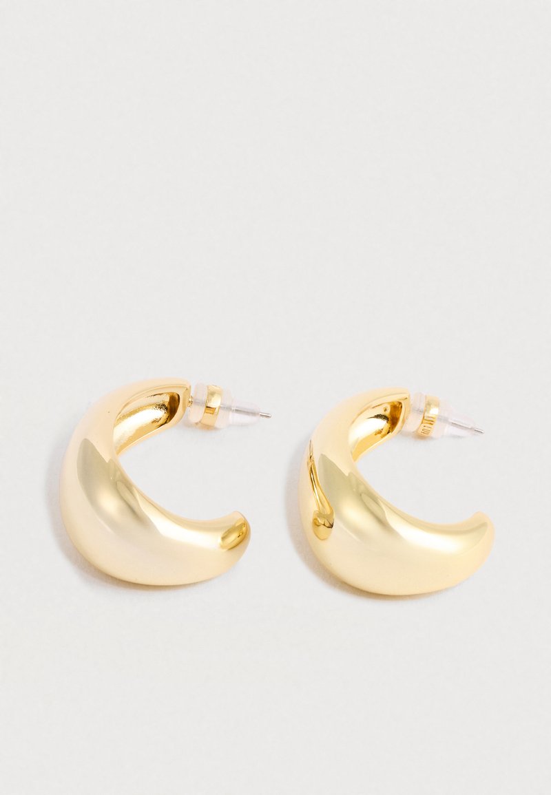 Une paire de créoles en or brillant et épais en forme de croissant, avec des fermoirs transparents à pression sur un fond blanc.