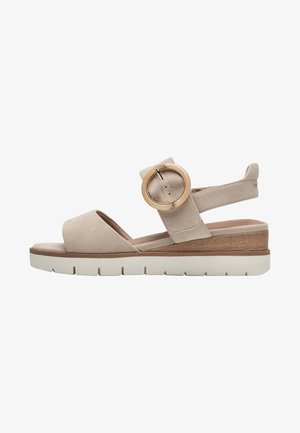 Beige suede wedge sandals with a wide toe and ankle strap, featuring a large circular buckle and wooden accent on a white sole.