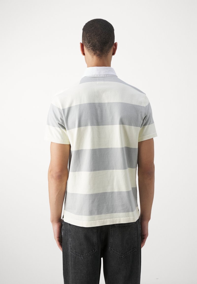 Homme portant un polo à manches courtes avec de larges rayures horizontales grises et écru, se tenant de dos, associé à un jeans noir.