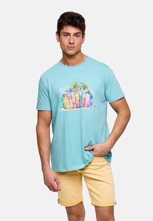 Hombre con camiseta azul claro estampada con tablas de surf coloridas y palmeras, combinada con pantalones cortos amarillos remangados, de pie contra un fondo blanco.