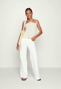 Top bege sem mangas, calças largas brancas e uma mala amarela de palha. Modelo está em uma postura descontraída e usa sandálias de salto pretas.