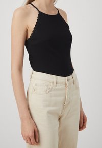 Débardeur noir à côtes avec des bords festonnés, assorti à un jean beige clair taille haute avec poches avant et fermeture à bouton métallique.