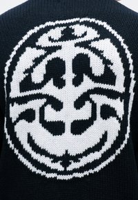 Čiernobiela pletená emblema na látke, s asymetrickým abstraktným dizajnom v kruhovej obrube.