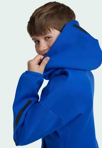 Blå huvtröja med en mjuk textur, med en svart detalj på ärmen och en avslappnad passform, bärs av ett barn.