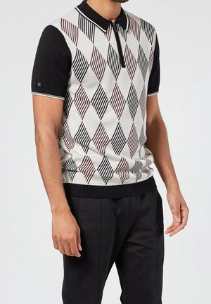 Man met een poloshirt met korte mouwen, zwarte mouwen en een beige diamantpatroon aan de voorkant, gecombineerd met een zwarte broek, staand tegen een effen achtergrond.