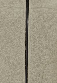 Veste en polaire beige clair avec une fermeture éclair noire, un tissu texturé et des détails cousus le long des coutures et des poches.