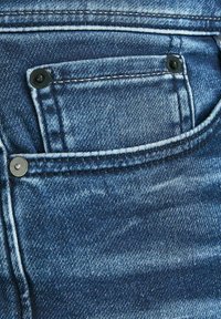 Modré denimové džíny mají malou přední kapsu se dvěma černými kovovými nýty a kontrastním šitím, které ukazuje na vybledlou strukturovanou látku.