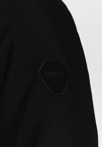 Jaqueta de tecido preto com um patch hexagonal em couro preto com "ONLY" em relevo. Textura suave e design simples.