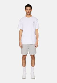 Vit t-shirt med kort ärm och en liten svart logotyp, i kombination med grå shorts. Modellen bär vita strumpor och sneakers mot en enfärgad bakgrund.