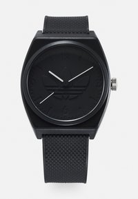 Montre analogique noire avec un boîtier carré, un bracelet texturé noir et un logo en relief sur une lunette mate. Les aiguilles des heures et des minutes blanches contrastent avec le cadran.