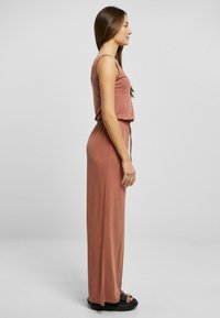 Ribbet, ermeløs jumpsuit i en dempet terrakottafarge. Har en justerbar snor i midjen og når til ankelen. Flate sandaler brukes.