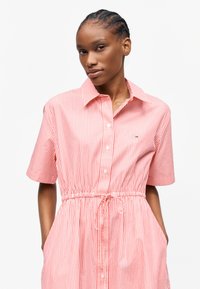 Γυναίκα με πλεξούδες που φοράει ροζ και λευκό φόρεμα με κάθετες ρίγες, κοντά μανίκια, κορδόνι στη μέση και κουμπιά μπροστά.