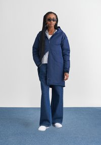 Giubbotto imbottito blu navy con cappuccio, tasca frontale e zip; abbinato a pantaloni in denim a gamba larga e scarpe da ginnastica bianche. Tessuto strutturato.