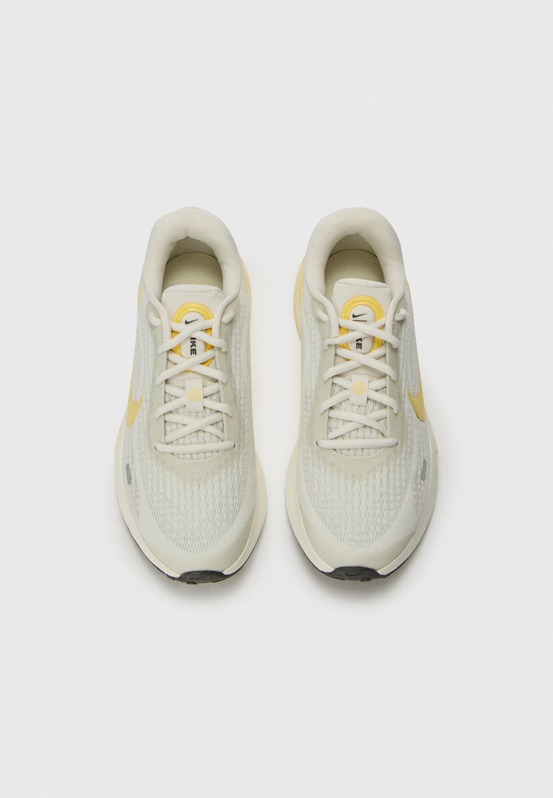 Baskets de sport beige clair avec tige en maille texturée, accents jaunes, semelle rembourrée et laçage traditionnel. Logo présent sur la langue.