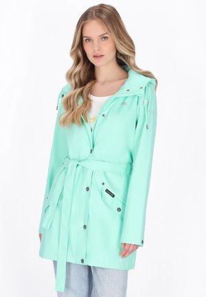 Kobieta z długimi blond włosami, ubrana w miętowozielony płaszcz przeciwdeszczowy z paskiem, na białą koszulę i jasnoniebieskie dżinsy, stojąca na białym tle.