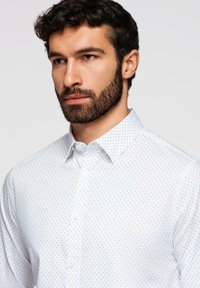 Homme aux cheveux bouclés foncés et barbe portant une chemise blanche à boutons avec un petit motif de points foncés, regardant légèrement sur le côté.