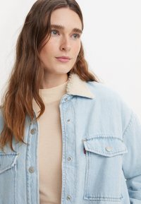 Lyseblå denimjakke med falsk pelskrage, trykknapper og brystlomme. Stoffet har en glatt tekstur og et uformelt design.