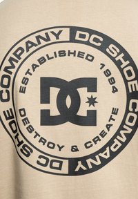 Beige bavlněný svetr s výrazným černým kulatým logem obsahujícím texty "DC Shoe Company," "Založeno 1994" a "Destroy & Create."