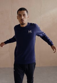 Felpa blu navy con scollatura rotonda e polsini a costine, con il logo bianco di Under Armour sul petto. Indossata con pantaloni neri.