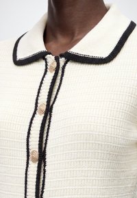 Haut en maille crème avec bordure noire le long du col et de la ligne de boutons, avec des boutons décoratifs dorés texturés.