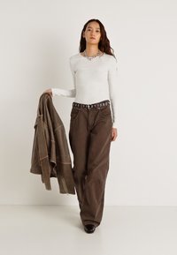 Šviesiai pilkas ilgomis rankovėmis megztinis, rudos spalvos plačios kelnės su diržu, kuriame yra apskritimų formos iškirpimai, ir rudą striukę laiko rankoje.