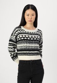 Adīts džemperis krēmkrāsā un melnā, ar ģeometriskiem rakstiem, platu ribota apakšmalu un apaļu izgriezumu. Izgatavots no teksturētas dzijas.