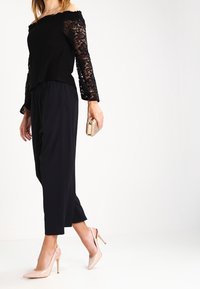 Top nero senza spalle con maniche in pizzo, abbinato a pantaloni neri a vita alta e scarpe con tacco nude; tiene in mano una borsa a tracolla dorata.