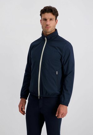 Man met kort donker haar, gekleed in een marineblauwe jas met ritssluiting en elastische manchetten, en een marineblauwe broek, staand tegen een effen lichte achtergrond.