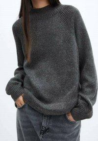 Maglione grigio a maglia con collo alto a coste, dal fit ampio e polsini a coste. Indossato sopra jeans in denim scuro, presenta una morbida texture e uno stile casual.