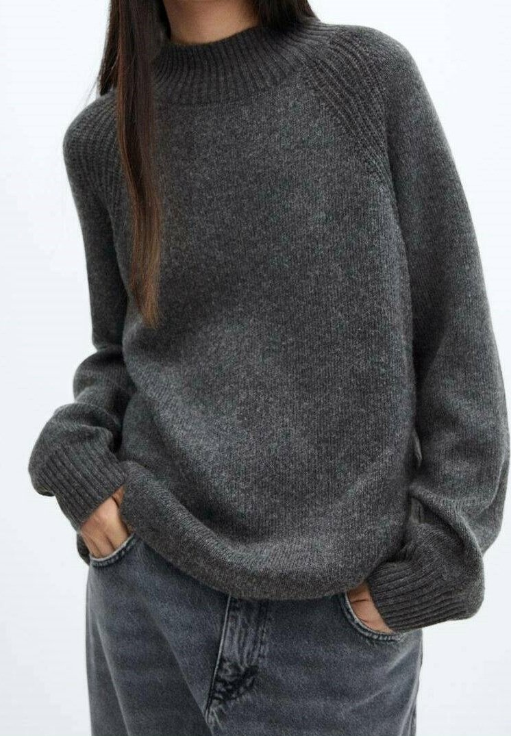 Maglione grigio a maglia con collo alto a coste, dal fit ampio e polsini a coste. Indossato sopra jeans in denim scuro, presenta una morbida texture e uno stile casual.