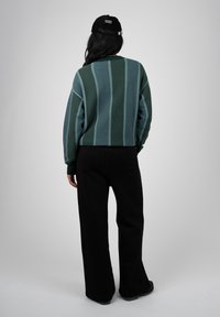 Personne debout de dos, portant un pull rayé vert et bleu, un pantalon noir et une casquette noire sur un fond gris uni.