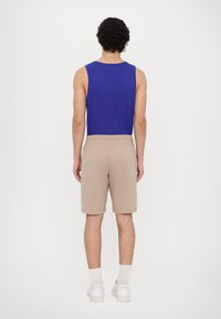 Blå sans ermer skjorte og beige shorts med elastisk midje. Modellen har på seg hvite joggesko og ankellange sokker, sett fra baksiden.