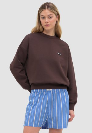 Junge Frau trägt ein dunkelbraunes Sweatshirt und blau-weiß vertikal gestreifte Shorts, steht vor einem einfarbigen Hintergrund.