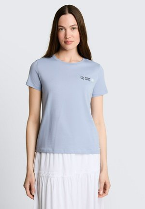 Femme aux longs cheveux bruns portant un t-shirt bleu clair avec le texte "You Make My Daisy" et une petite illustration de marguerite, associée à une jupe blanche.