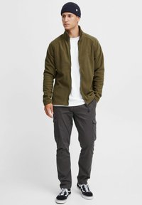 Olivgrüne Fleecejacke mit hohem Kragen und Reißverschluss, kombiniert mit dunklen Cargo-Hosen und schwarzen Sneakern. Leicht und lässig geschnitten.